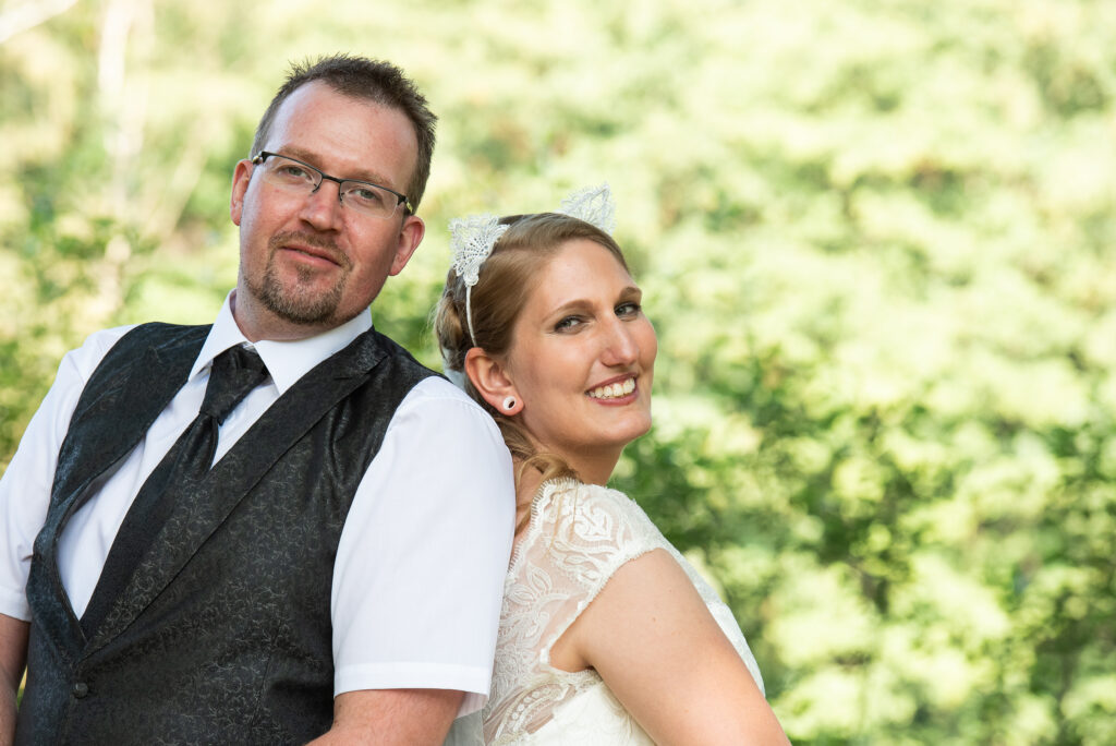 Das Brautpaar steht Rücken an Rücken vor einen unscharfen grünen Hintergrund uns strahlt in die Kamera
Hochzeitsfotografen
