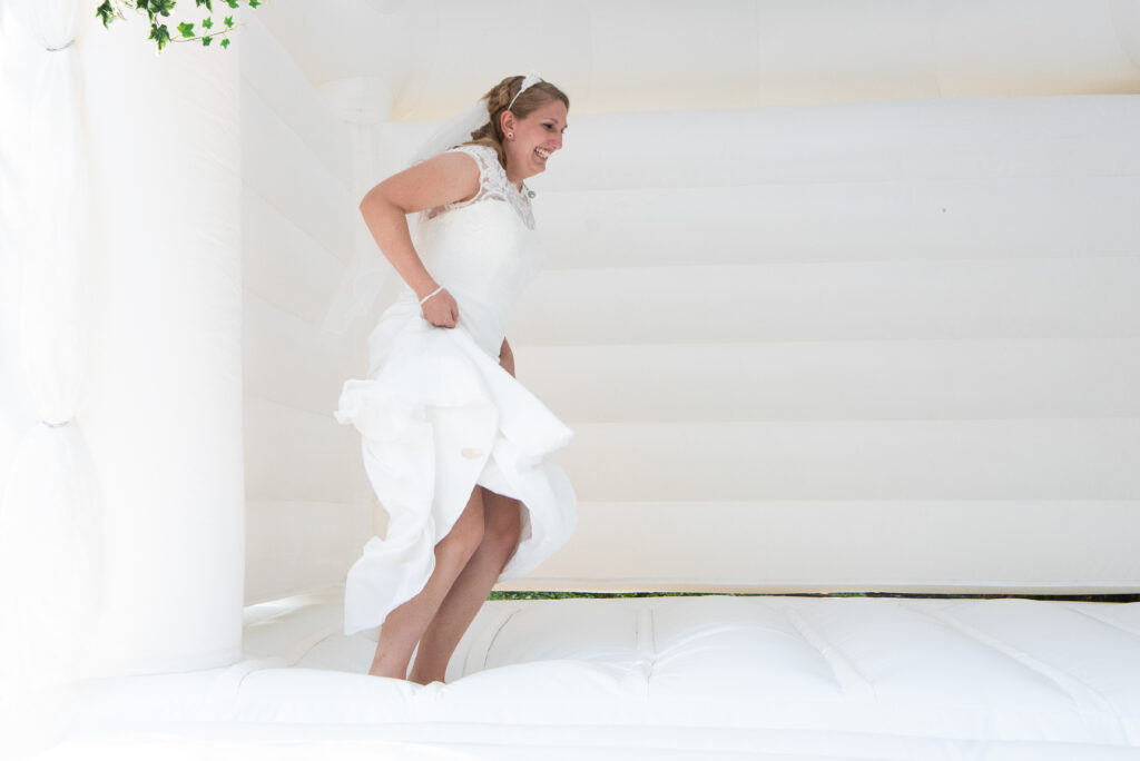 Eine weiße Hüpfburg für die Braut.
Die Braut hebt hier Kleid mit beiden Händen hoch uns springt.
Hochzeitsfotografen