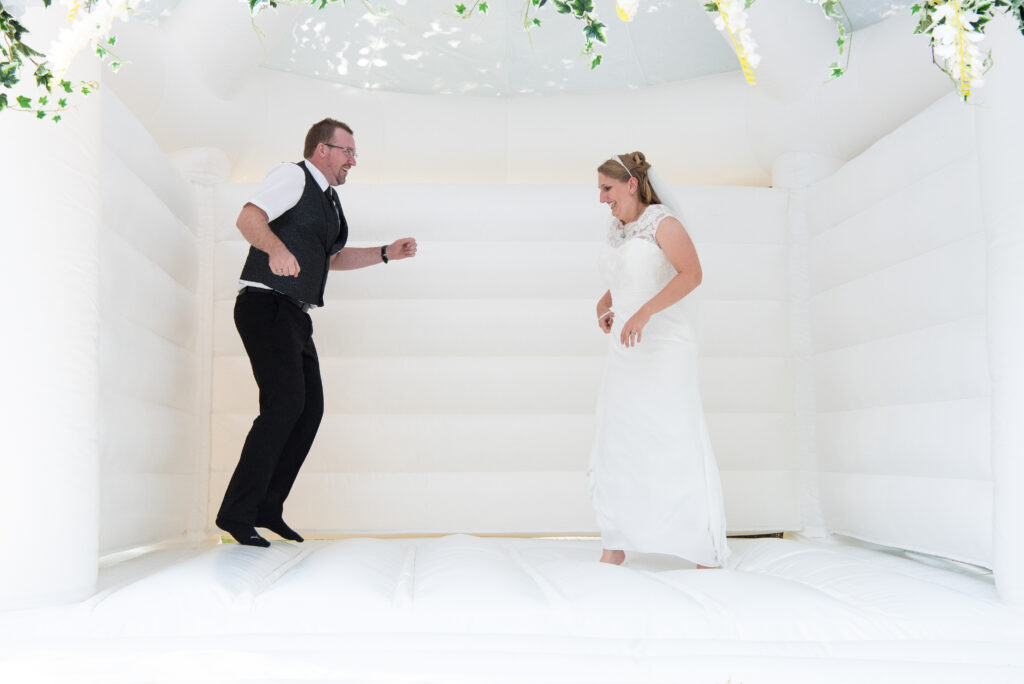 Der Bräutigam springt hoch und die Braut schaut zu, wie ihr Mann in der weißen Hüpfburg hüpft
Hochzeitsfotografen